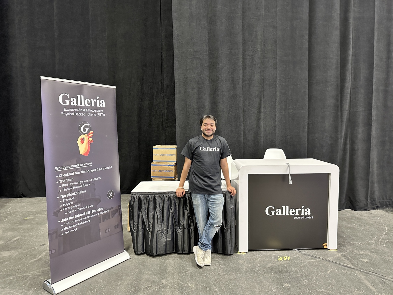 JC standing at the Gallería booth at NFT.NYC: a banner, a branded counter, and demo table